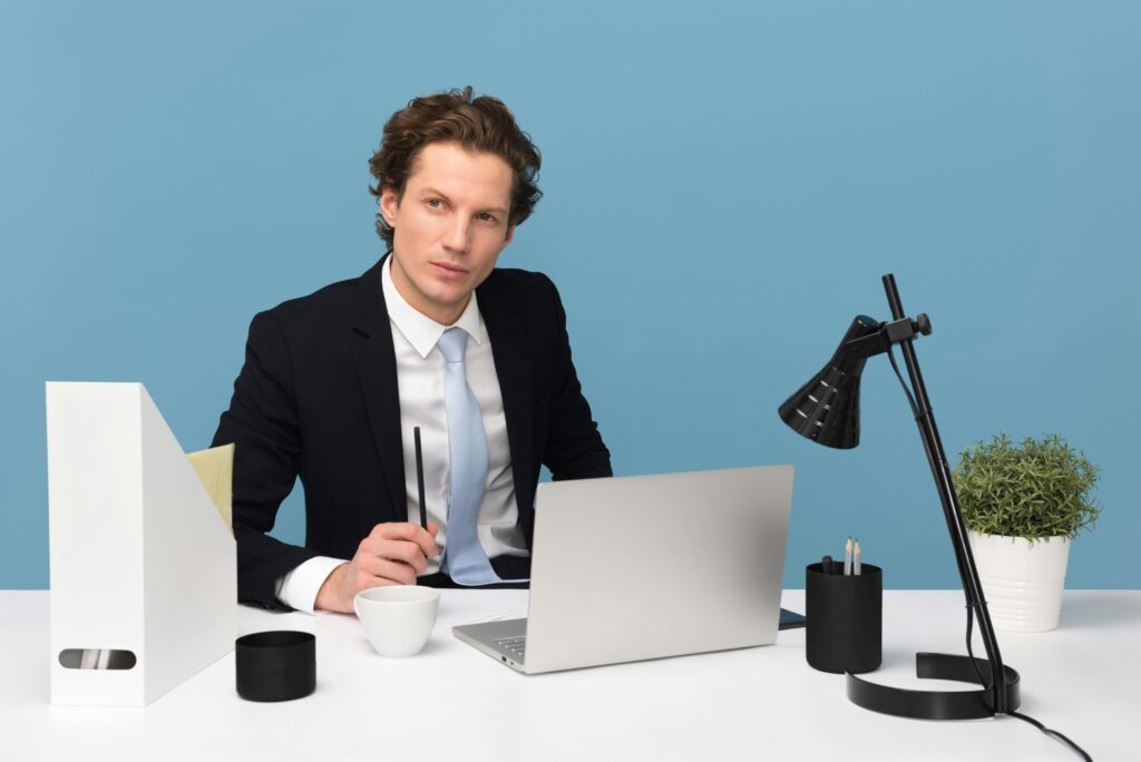 A focused businessman in a suit works at his tidy desk with a laptop, plant, and lamp.